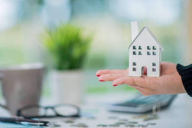 A hand holds out a 3D model of a house in this real estate concept photo.  A calculator, loose change, glasses, coffee mug and a play lay out on a table in the background.